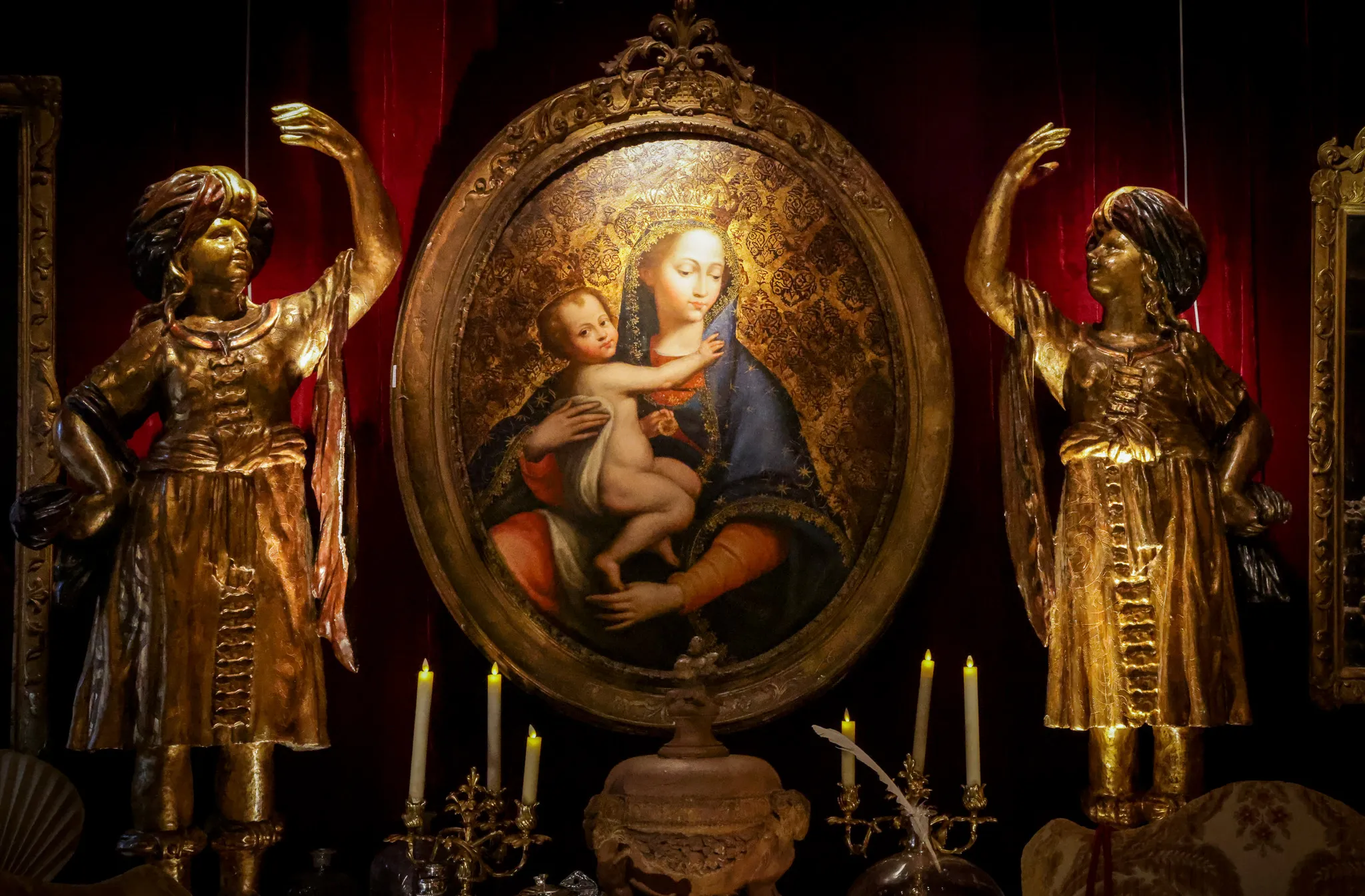 Tableau ovale de la Vierge à l'Enfant encadré par deux statues dorées sur fond rouge.
