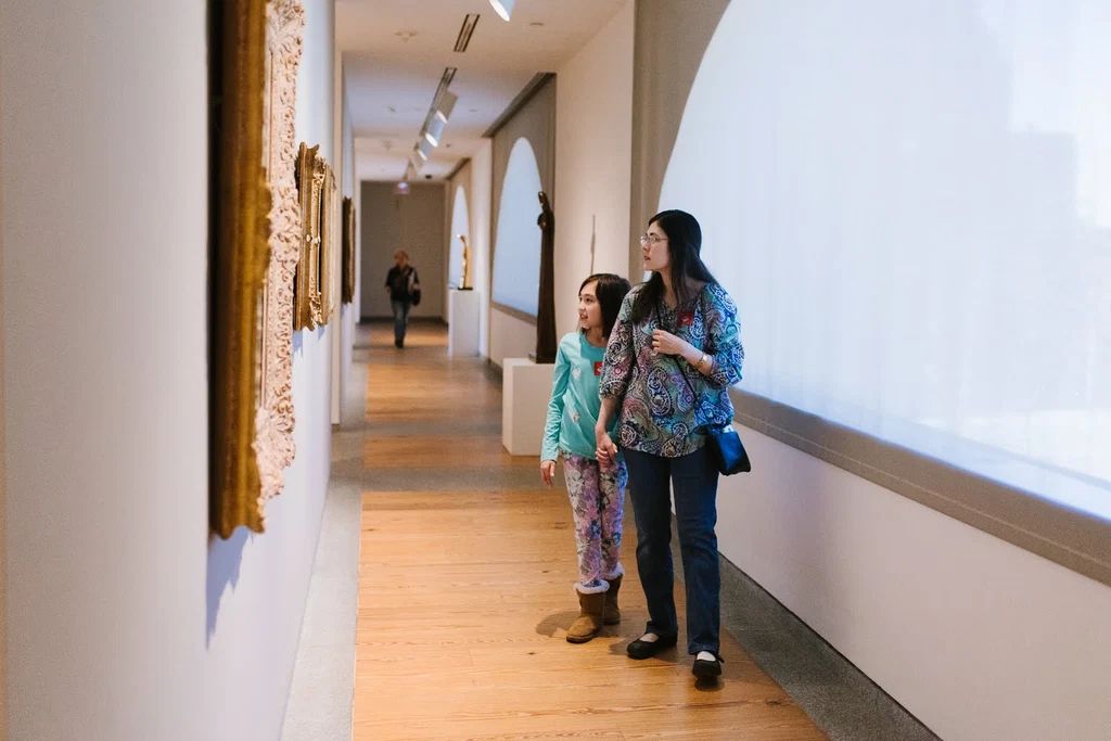 A parent and child walking a Portland Museum of Art gallery, looking at framed works along the wall