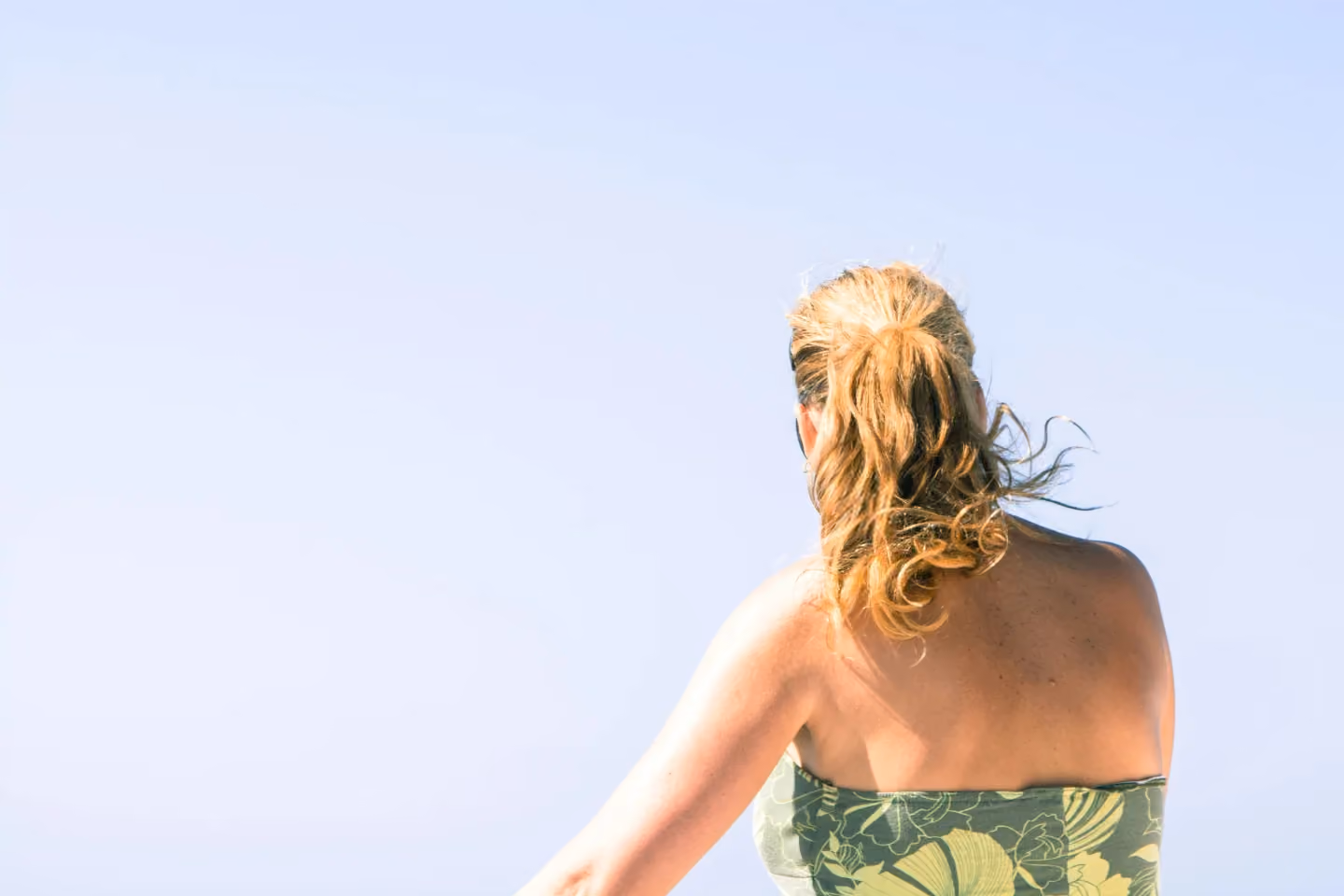 Woman with blonde hair tied in a ponytail wearing a green patterned strapless top, standing outdoors with her back to the camera against a clear blue sky.