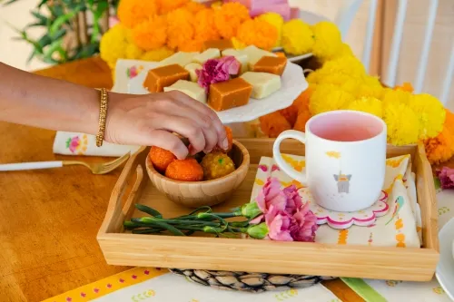 A hand reaches for Indian mittai sweets served in a platter with tea and marigold decor.