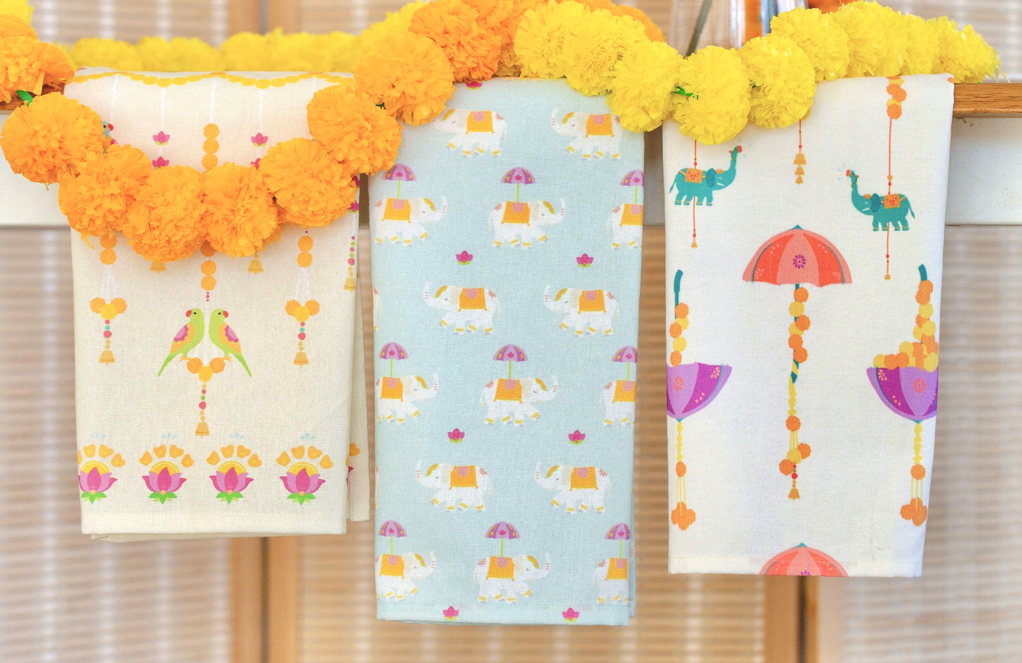 Trio of Diwali tea towels with elephants, parrots, and marigold motifs, draped with marigold garlands.