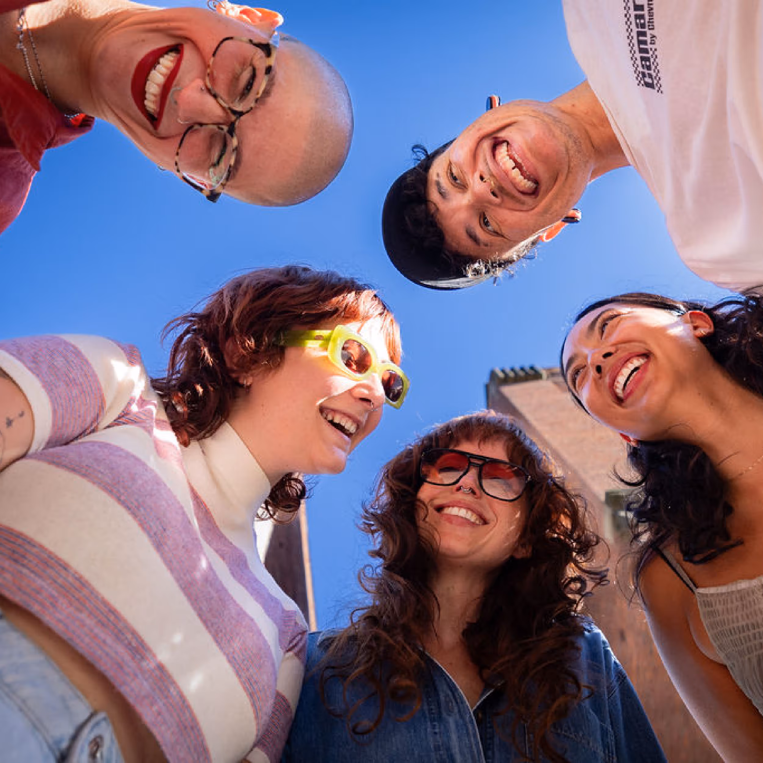 Five diverse friends smiling and leaning in a circle outdoors under a clear blue sky.
