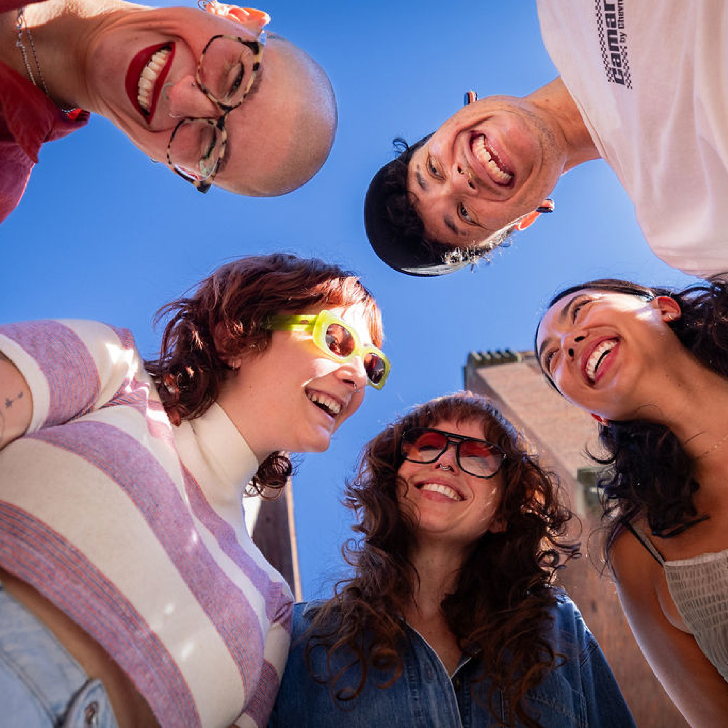 Five diverse friends smiling and leaning in a circle outdoors under a clear blue sky.