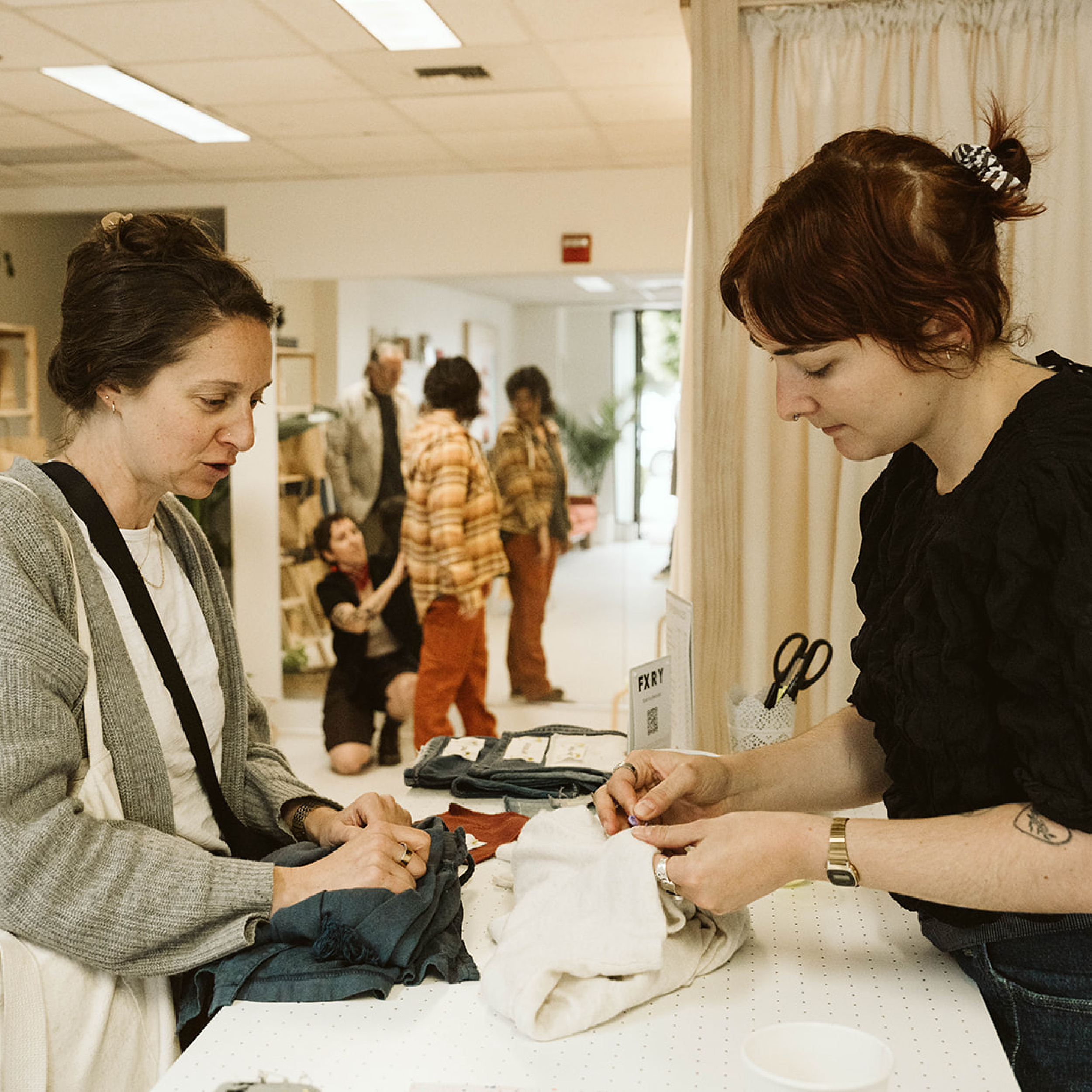 Two women folding clothes at a table in a bright indoor space with others in the background.