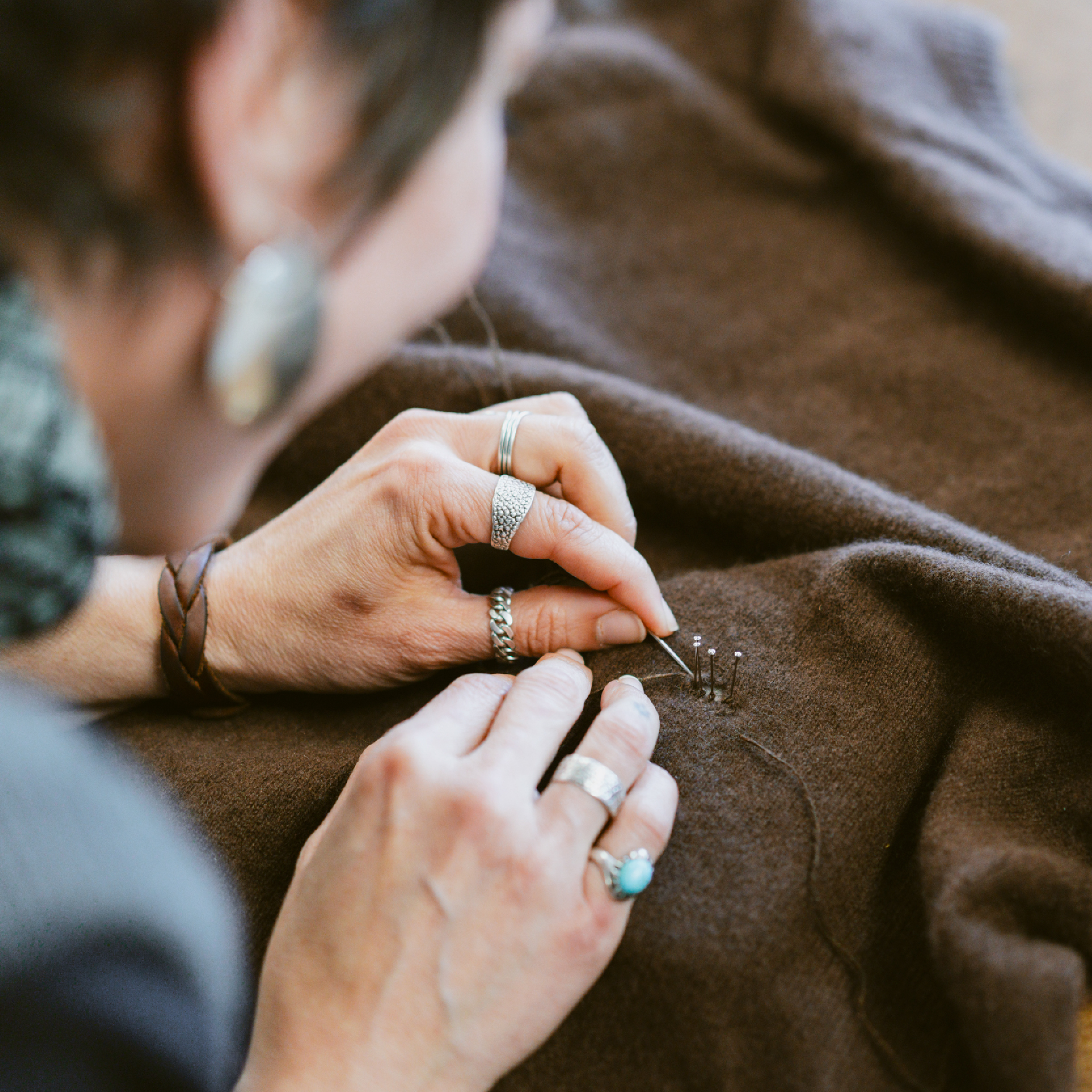 Person reknitting a brown sweater using a needle and pins, adorned with multiple rings and a bracelet.