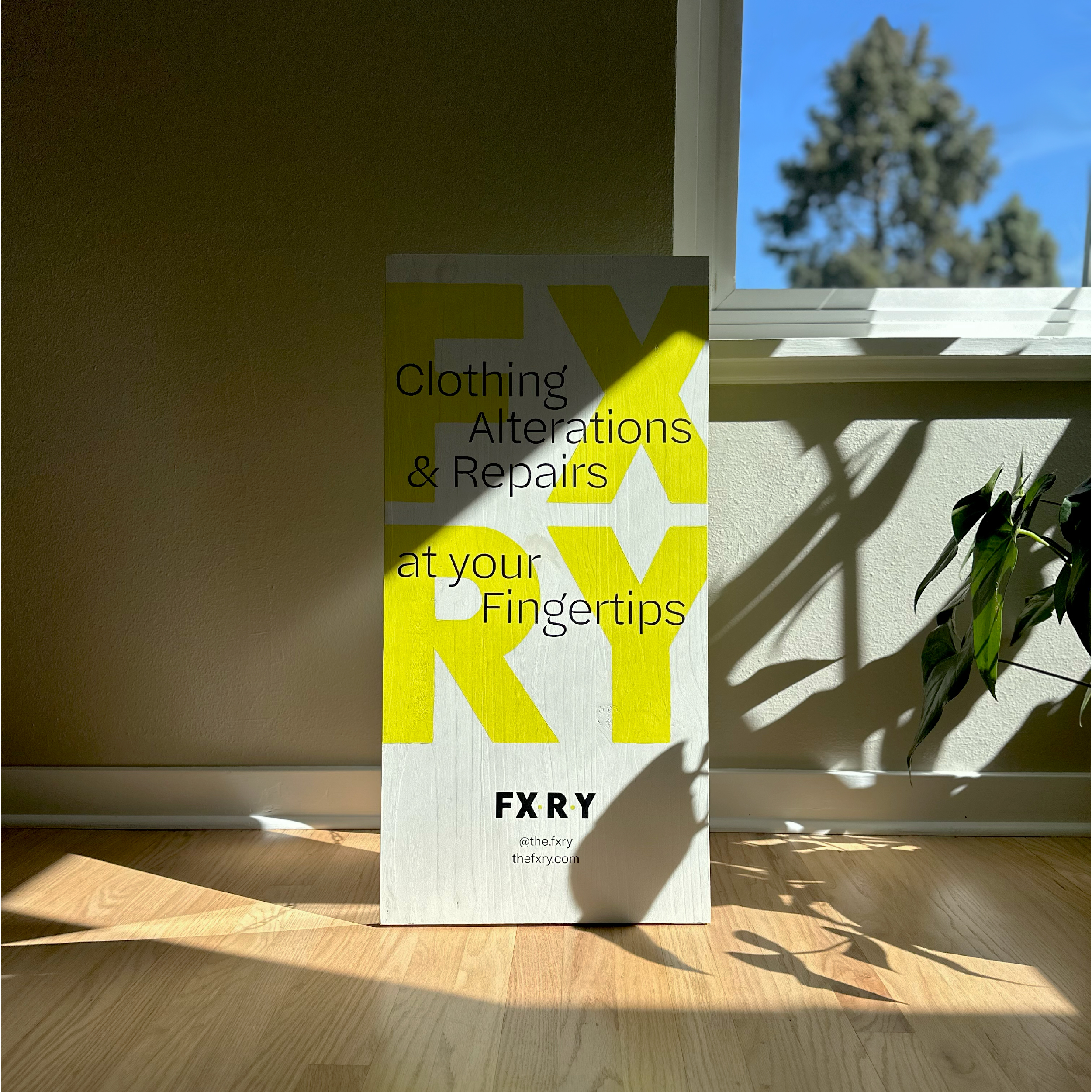 Signboard on a wooden floor by a wall with window shadows, displaying text for clothing alterations and repairs by FXRY.