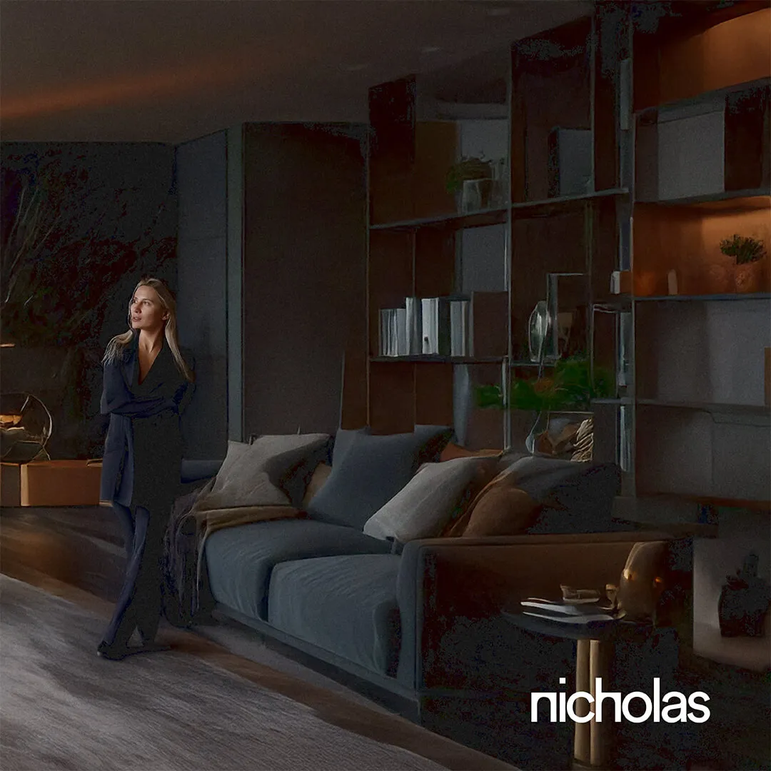 Dark grey luxury hotel suite with a bed, sofa, built-in bookshelf, and a woman standing in simple elegant pajamas, showcasing thoughtful, atmospheric interior design