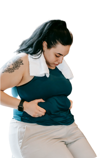 A woman with dark hair in a ponytail, wearing a dark green tank top and light-colored pants, stands looking down at her midsection. She holds her stomach with both hands, showing a concerned expression. A white towel is draped around her neck, and she has a tattoo on her left arm. The background is transparent, focusing on her expression and body language.