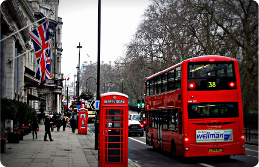 A vibrant city street in London, featuring iconic red elements: a double-decker bus, a telephone booth, and a mailbox. The Union Jack flag is prominently displayed on a building, and pedestrians walk along the sidewalk. Leafless trees line the street, adding a winter atmosphere. The bus route number "38" is visible on the bus.