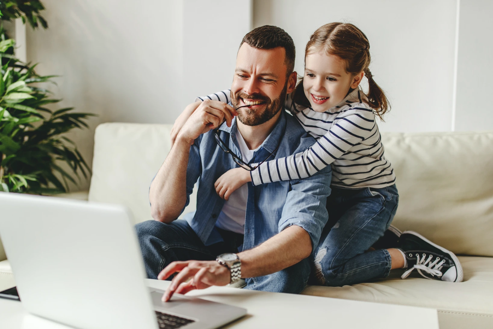 Adult male with brown hair and beard studying online while his young daughter hangs on his shoulders