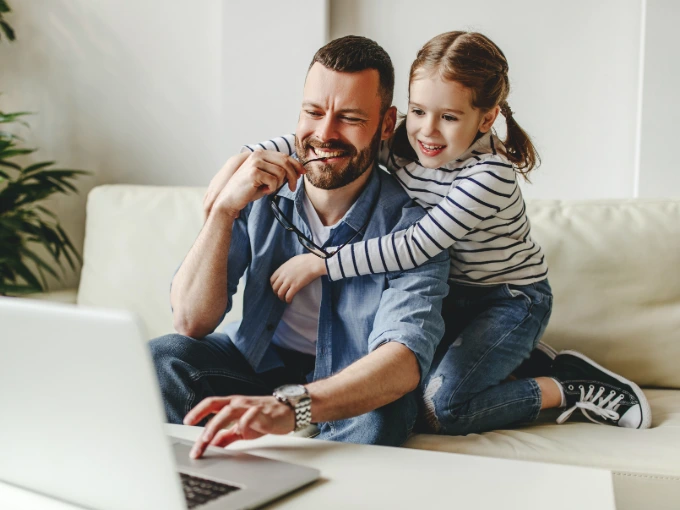 Adult male with brown hair and beard studying online while his young daughter hangs on his shoulders