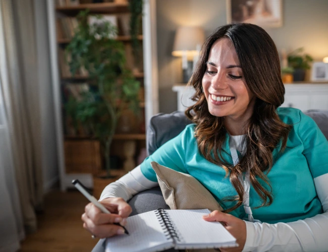 Female nurse in green scrubs sitting on a couch with planner open while conducting long-term goal setting for LPNs