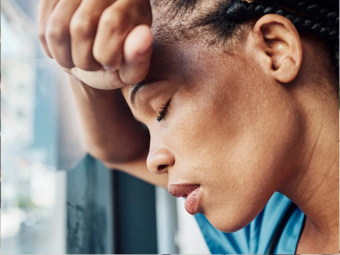 A nurse in scrubs resting her forehead on her arm by a window, appearing tired and overwhelmed.