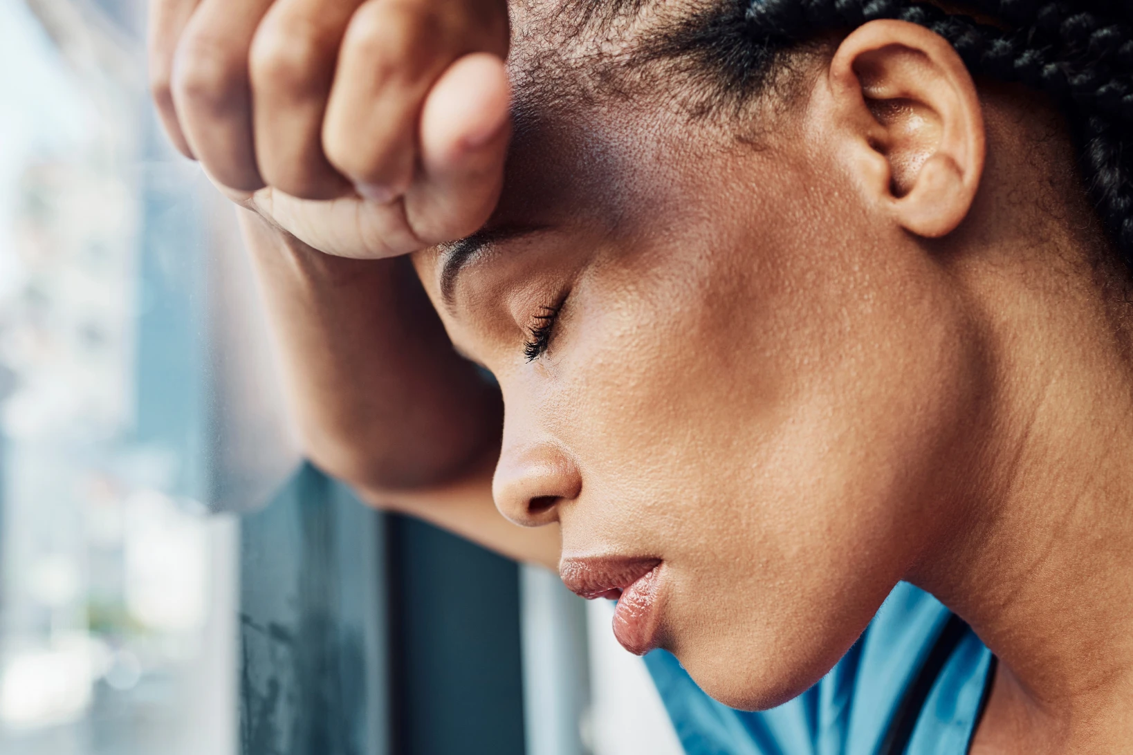 A nurse in scrubs resting her forehead on her arm by a window, appearing tired and overwhelmed.