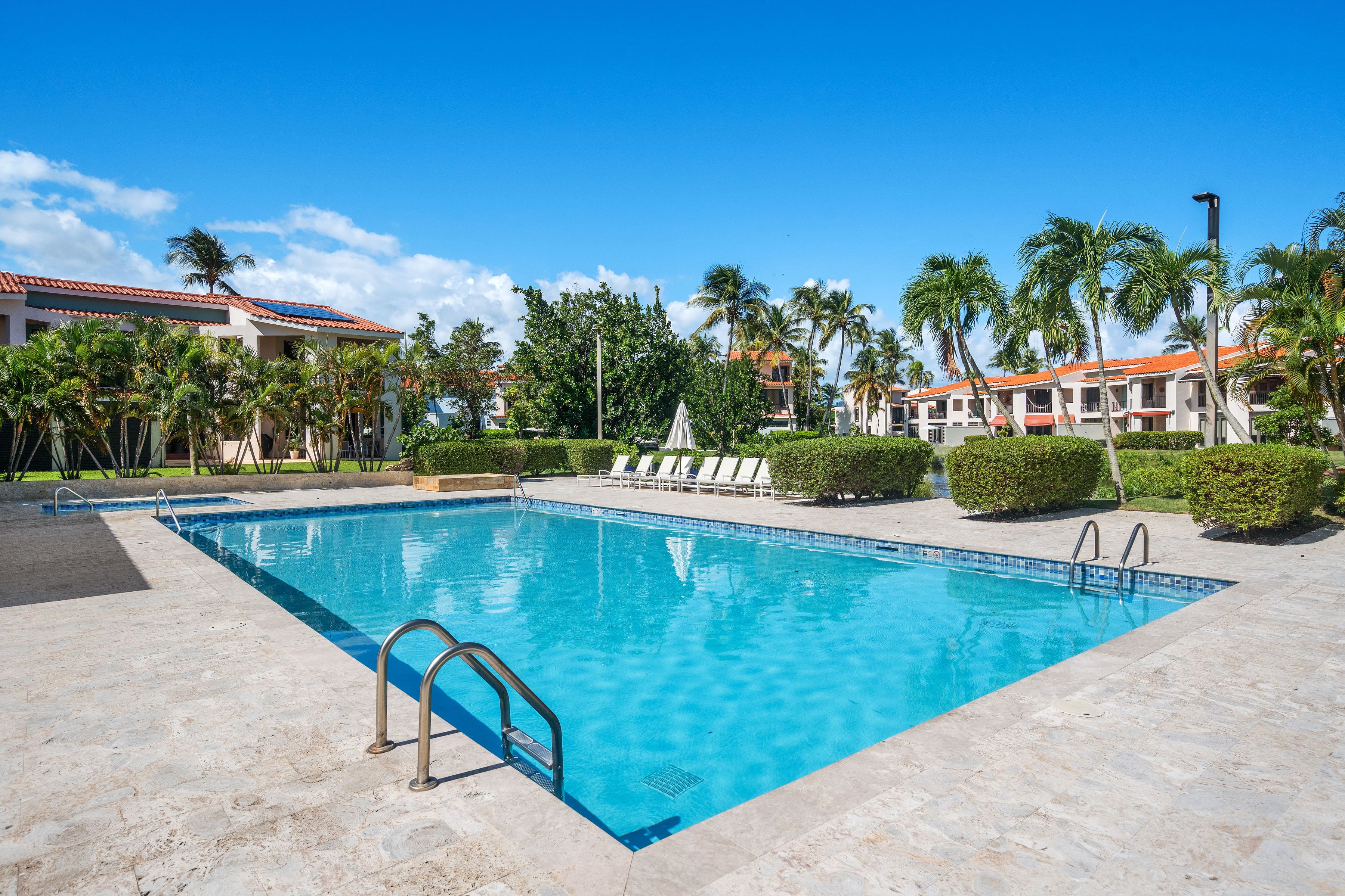 Large resort-style pool with bright blue water, lounge seating, palm trees, and Mediterranean-style buildings in the background at Fairlakes Village, Palmas del Mar.