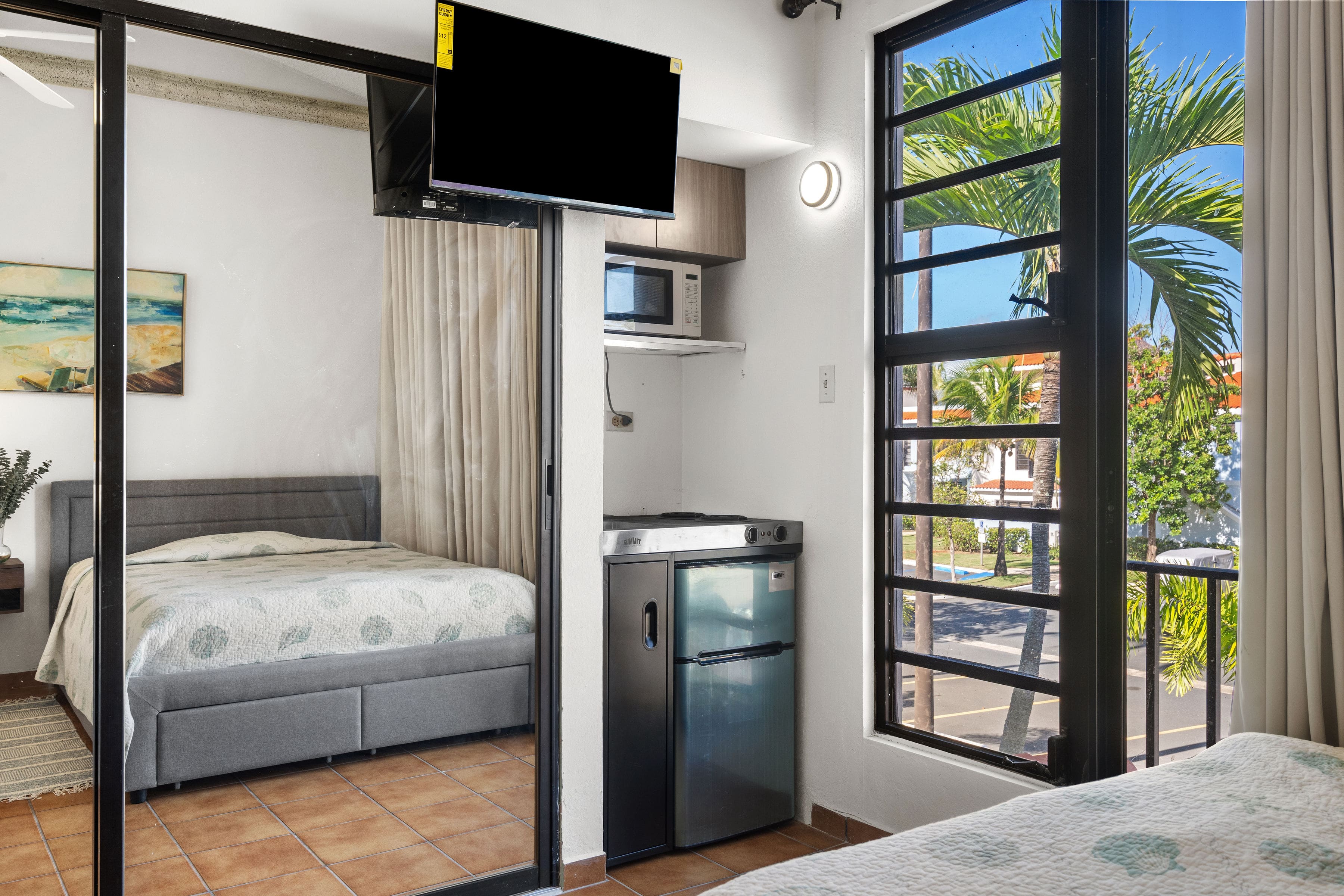 Guest suite kitchenette setup with mini-fridge, microwave, cooktop, wall-mounted TV, and large windows overlooking palm trees and the walkable Fairlakes Village neighborhood.