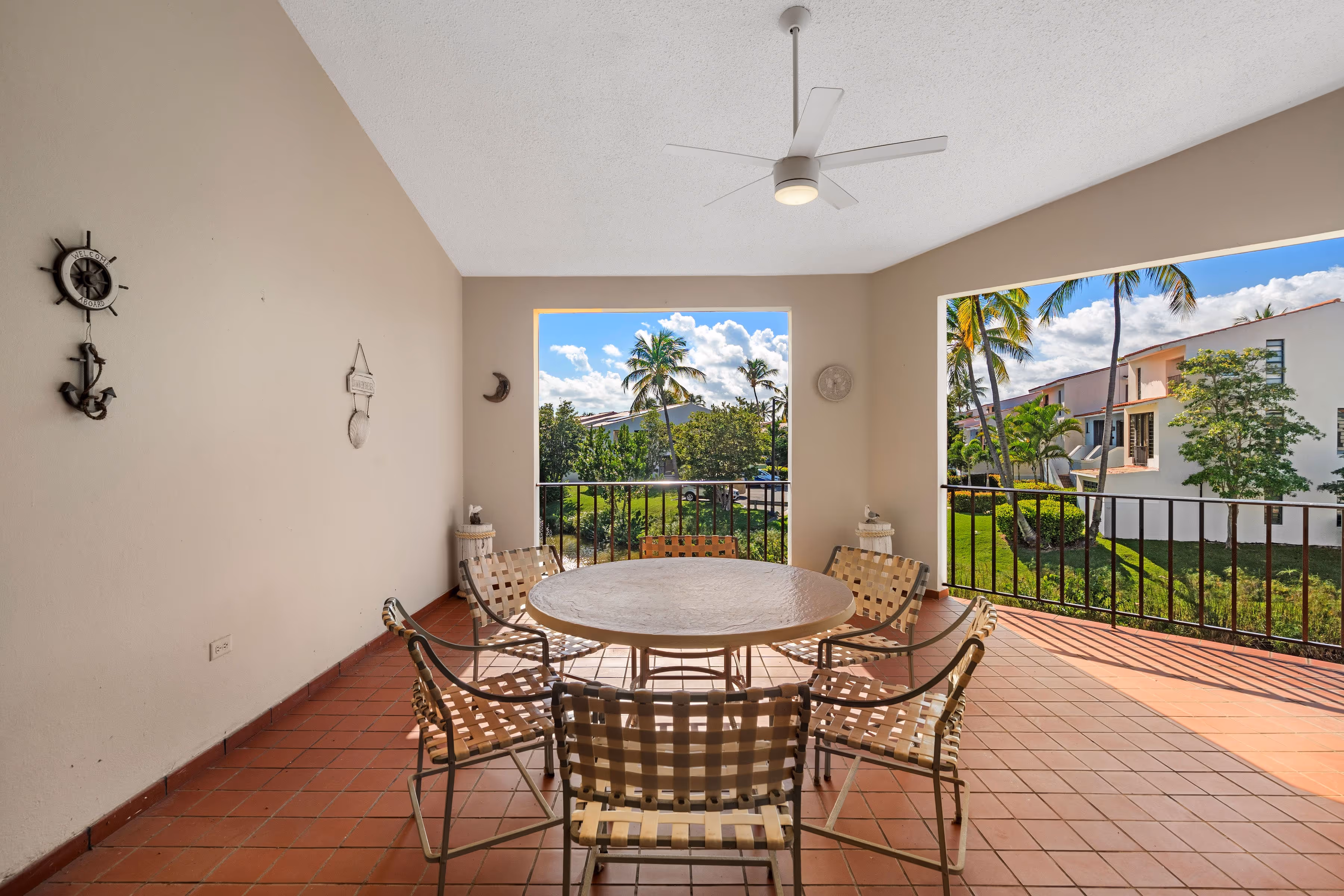 Covered outdoor terrace with round dining table, woven chairs, ceiling fan, and panoramic views of palm trees and the garden-style Fairlakes Village community.
