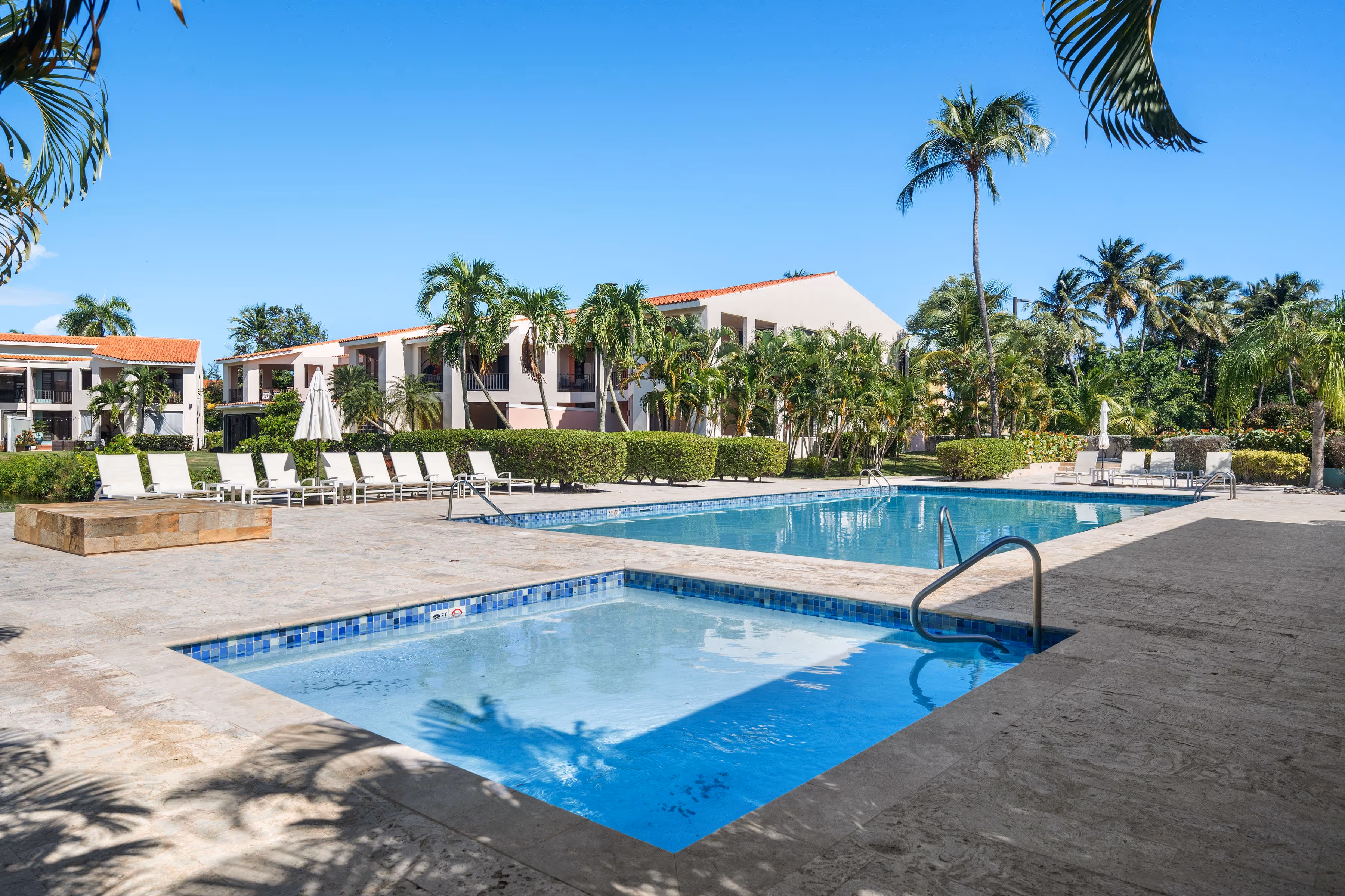 Fairlakes Village community pool area featuring a spacious main pool, children’s pool, sundeck loungers, tropical palm trees, and well-maintained garden surroundings.