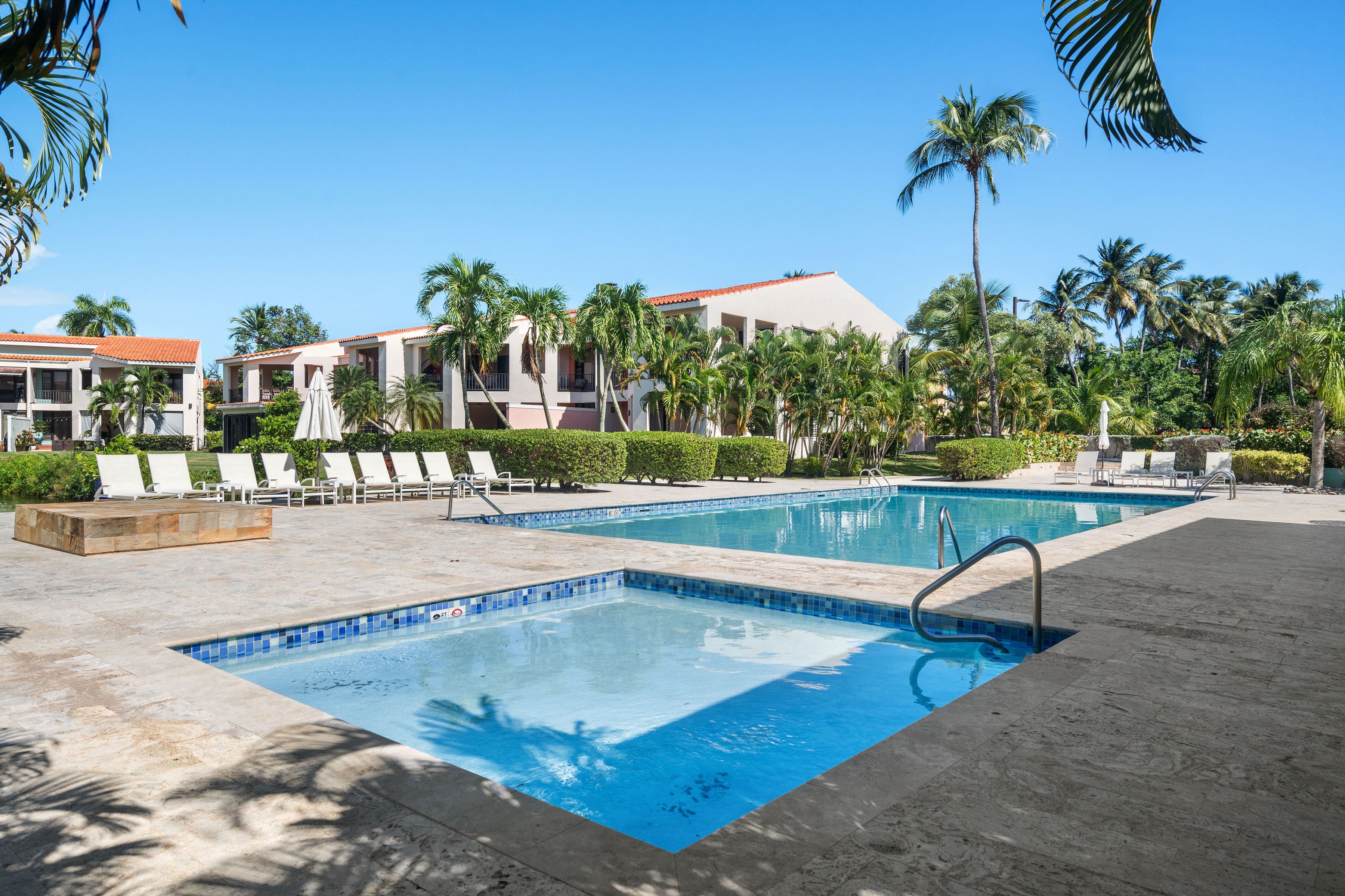 Fairlakes Village community pool area featuring a spacious main pool, children’s pool, sundeck loungers, tropical palm trees, and well-maintained garden surroundings.