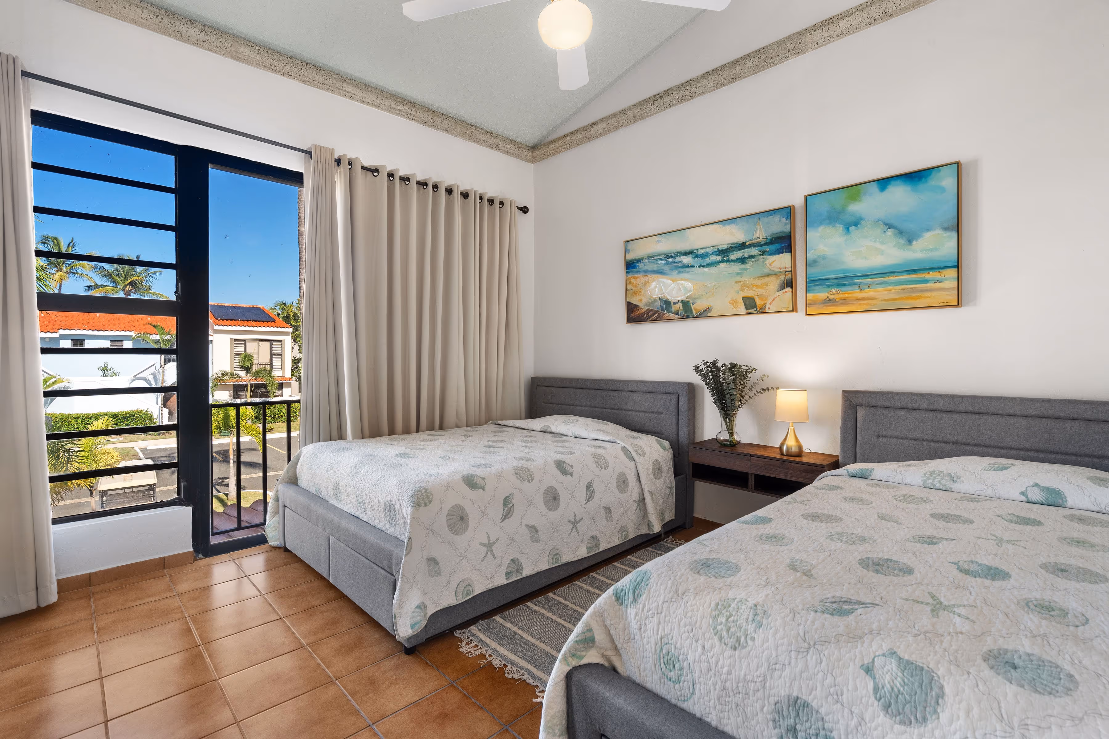 Guest bedroom with two beds, vaulted ceilings, beach-themed artwork, neutral curtains, and window views of the Fairlakes Village community and tropical landscaping.