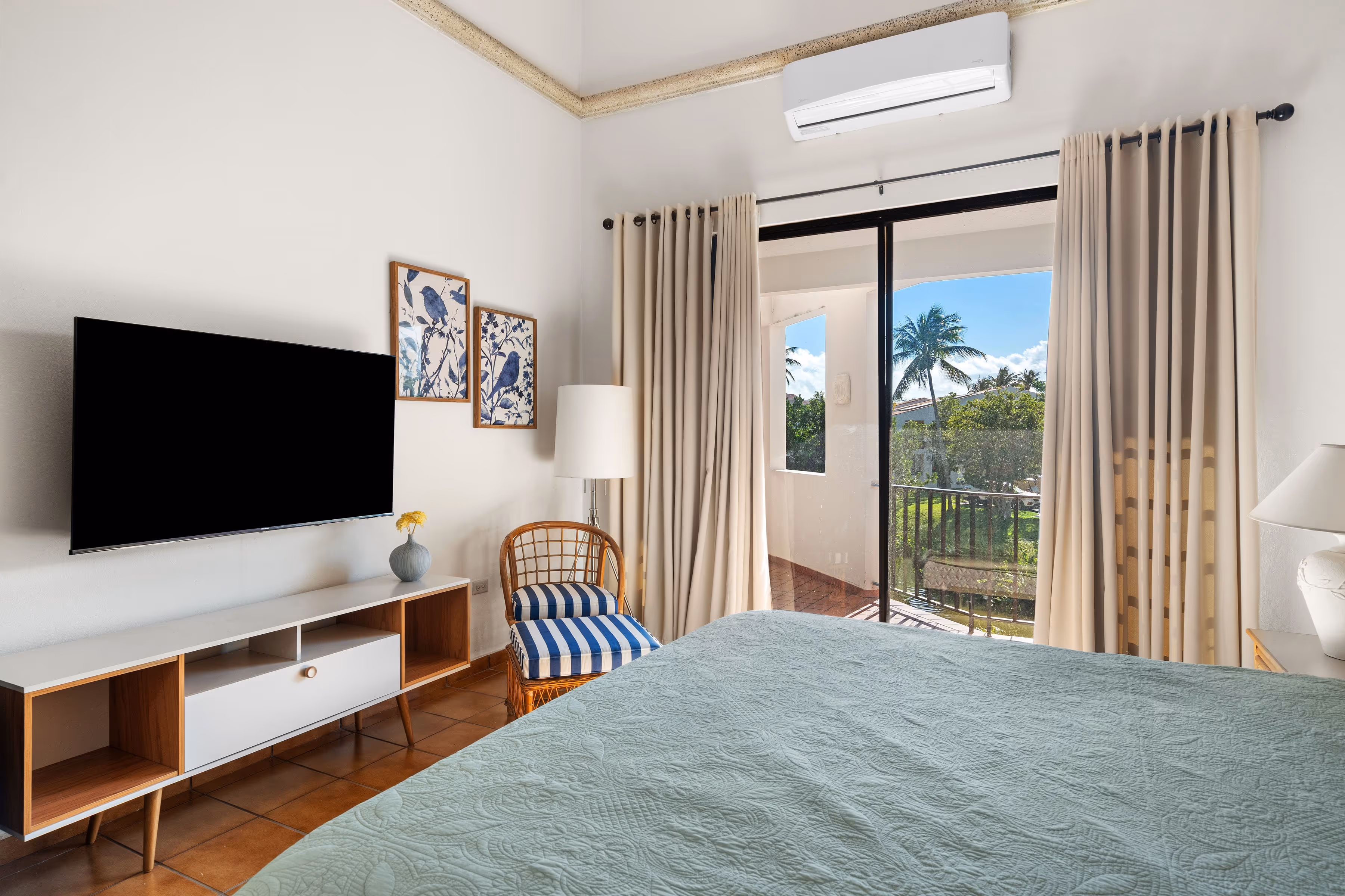Primary bedroom with sliding glass doors opening to a private terrace, featuring high ceilings, air conditioning, a mounted TV console, rattan chair with striped cushion, and views of tropical palms in Fairlakes Village, Palmas del Mar.
