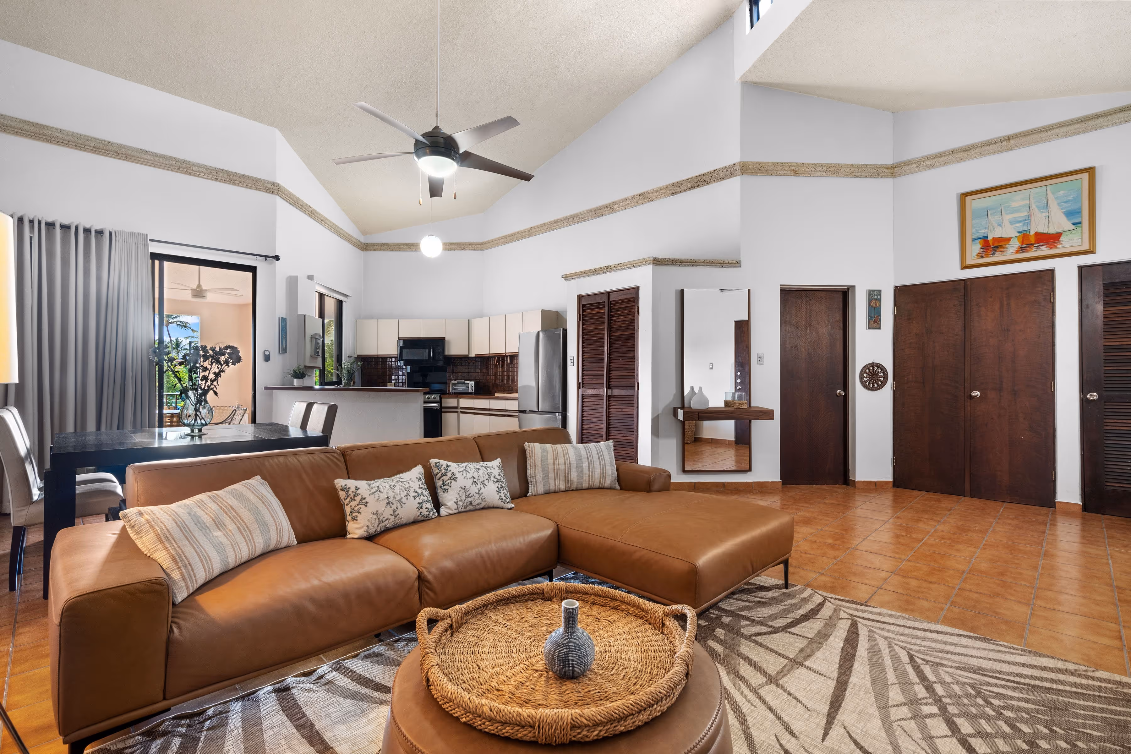 Living room and dining area layout showcasing a large leather sectional, tropical-themed rug, dining table for six, and seamless flow toward the kitchen and outdoor terrace in Palmas del Mar.