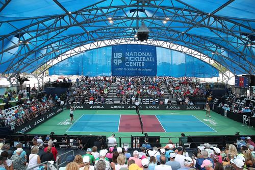 Indoor pickleball court with two players competing and a large audience seated under a blue canopy at the USOP National Pickleball Center.