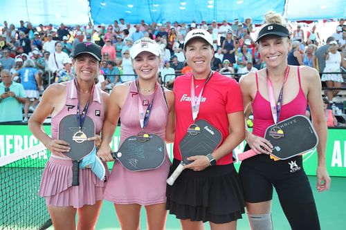 Four female pickleball players smiling and posing with paddles and medals on a court with a crowd in the background.