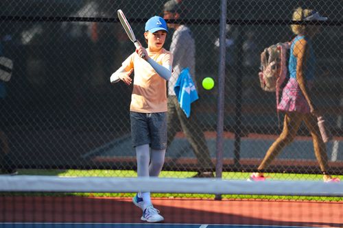Young boy in sportswear preparing to hit a tennis ball on an outdoor court with a chain-link fence in the background.