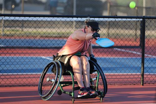 Woman in a wheelchair playing pickleball on an outdoor court, hitting a ball with a blue paddle.