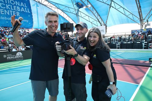 Three smiling adults on a pickleball court, one holding a microphone, another holding a professional camera on a tripod, and the third holding a smartphone.