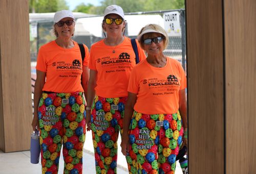 Three women wearing bright orange US Open Pickleball Championship shirts and colorful pants covered with multicolored pickleball illustrations and the phrase 'Eat Sleep Pickleball Repeat', standing and smiling outdoors near court 18.
