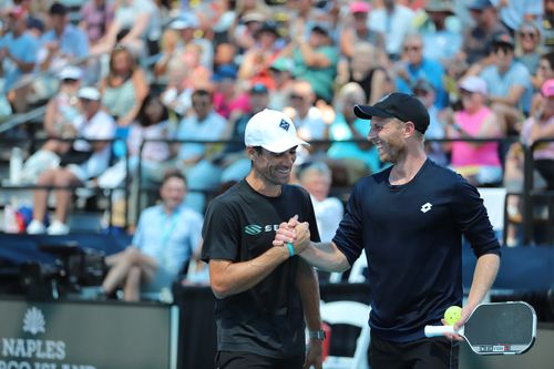 Two male pickleball players smiling and shaking hands on the court, one holding a paddle and ball, with a crowd in the background.