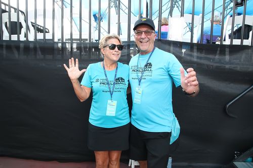 Smiling man and woman wearing matching blue US Open Pickleball Championship volunteer shirts posing with arms raised.