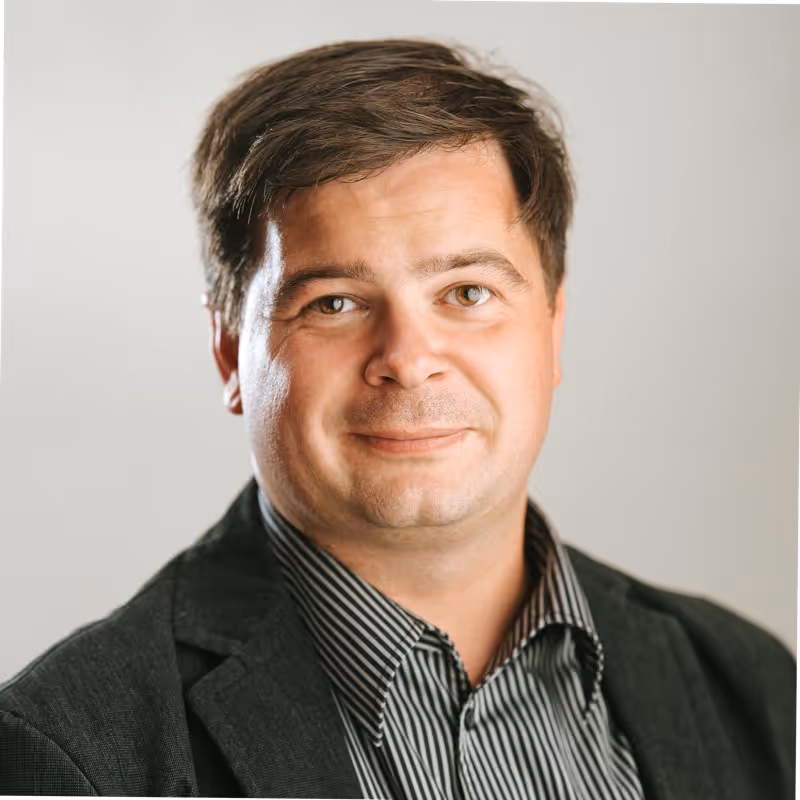 Smiling man with short brown hair wearing a dark blazer and striped shirt against a plain light background.