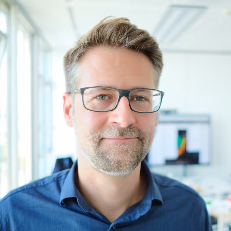 Smiling man with glasses and light brown hair wearing a blue shirt in a bright office setting.