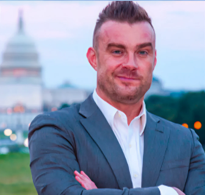 Man in a gray suit with white shirt standing with arms crossed in front of the U.S. Capitol building at dusk.