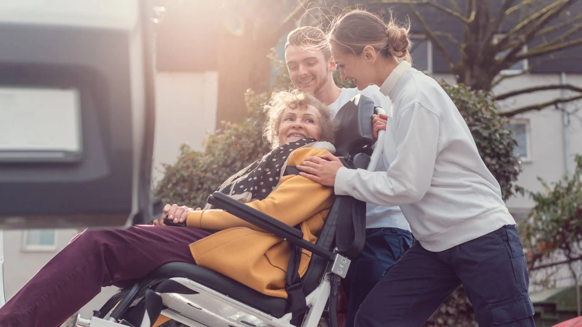 Two caregivers assist an elderly woman in a wheelchair outside, all smiling and interacting warmly.