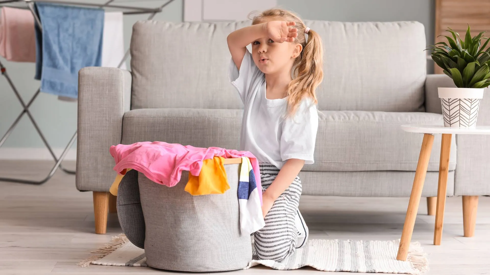 Little girl kneeling by laundry basket full of clothes, wiping forehead, in living room.