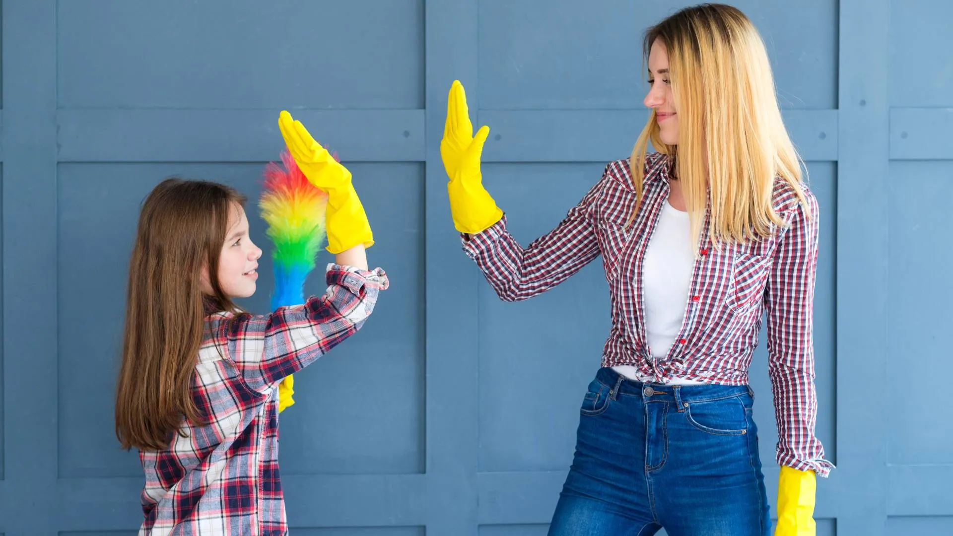 Woman and girl in yellow gloves high-five while holding a rainbow-colored duster in front of a blue wall.