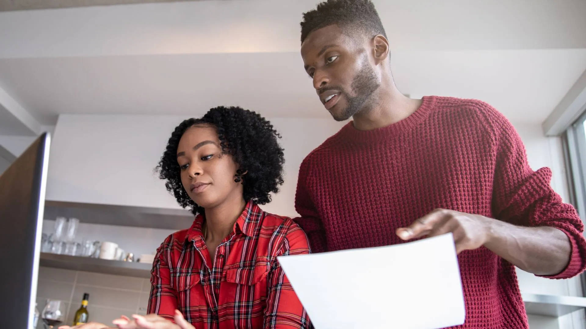 Couple in red shirts reviewing a document together while looking at a computer screen in a kitchen.
