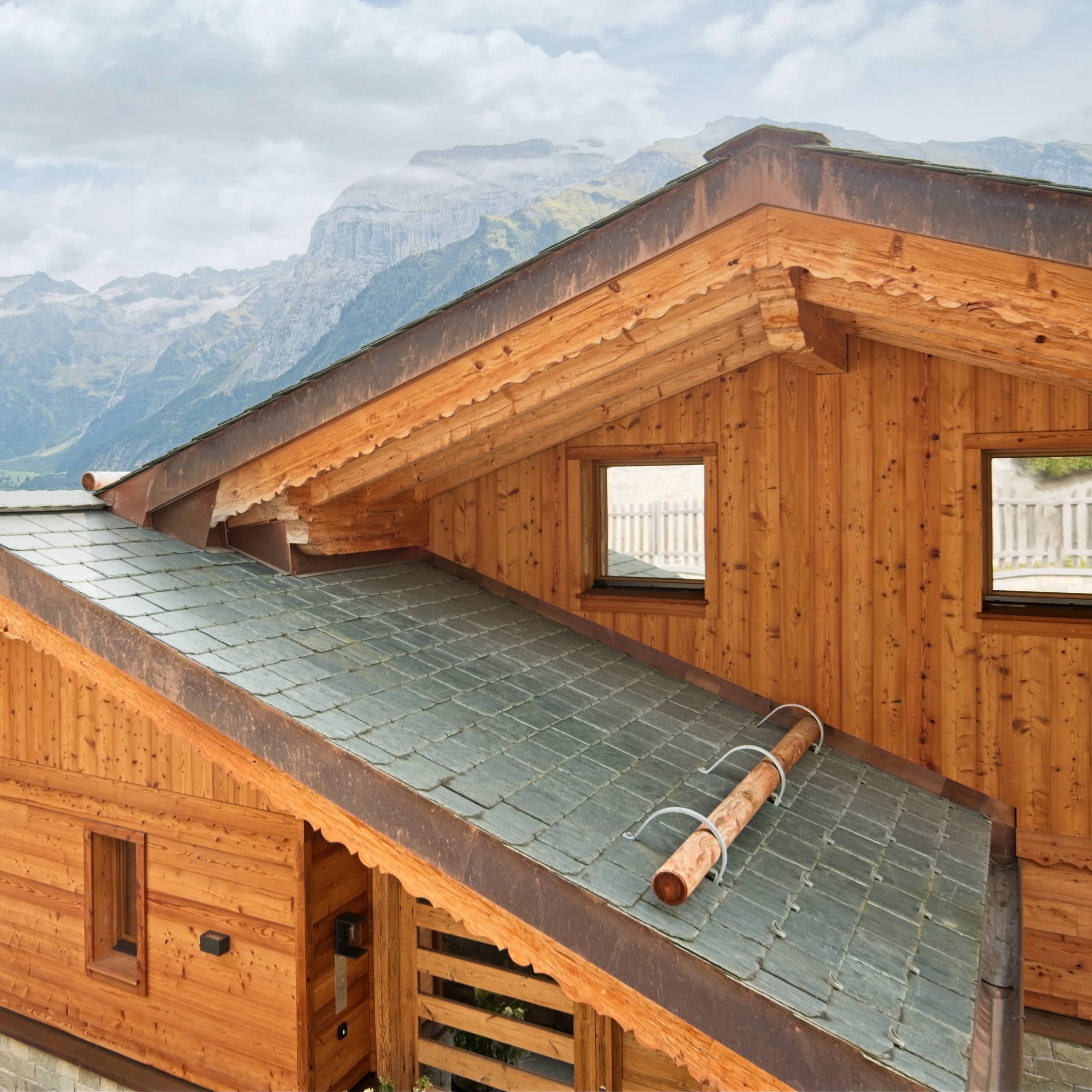 Dach mit Schieferplatten und Holzverkleidung eines Bergchalets vor bewölktem Bergpanorama.