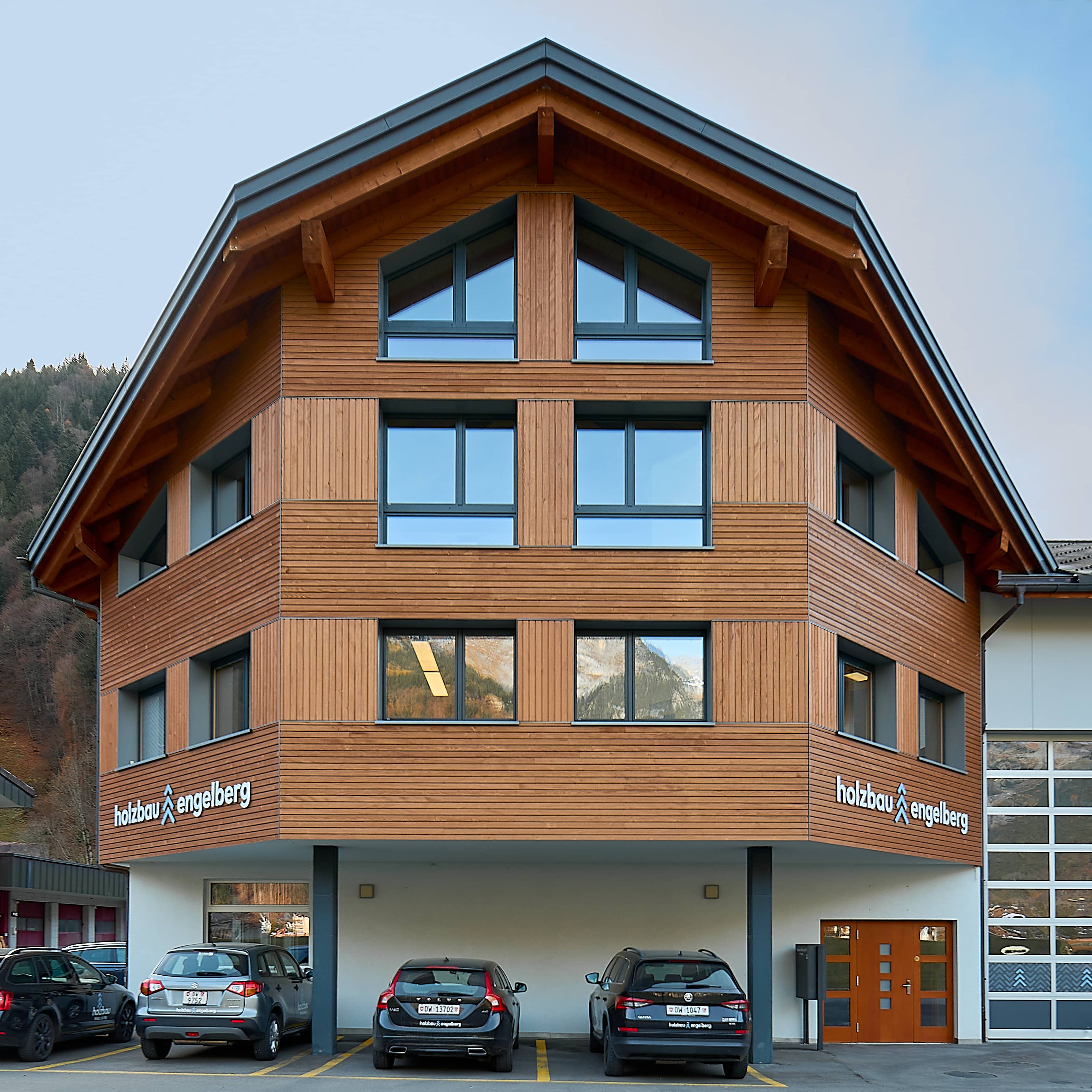 Mehrstöckiges Bürogebäude von Holzbau Engelberg mit Holzfassade und großen Fenstern, davor geparkte Autos und Berge im Hintergrund.