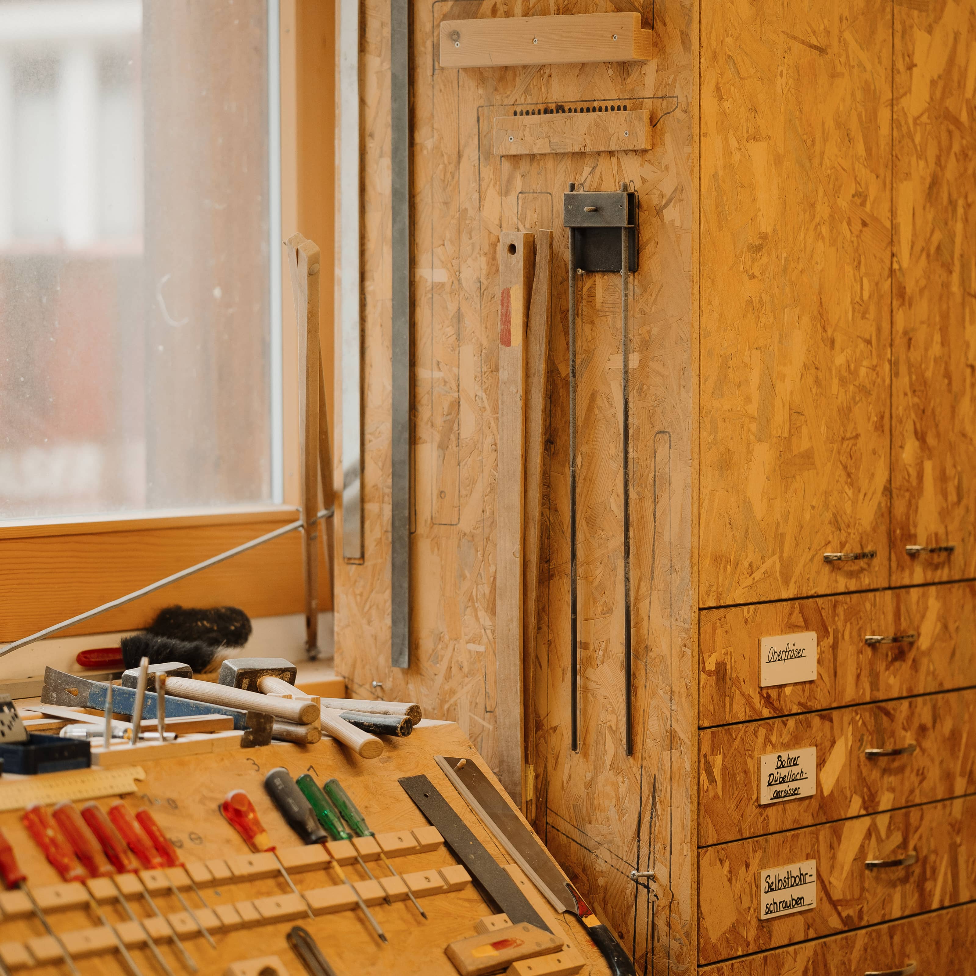 Werkbank mit verschiedenen Handwerkzeugen wie Schraubenziehern, Hämmern und Messwerkzeugen an einer Holzplatte vor einem Fenster.