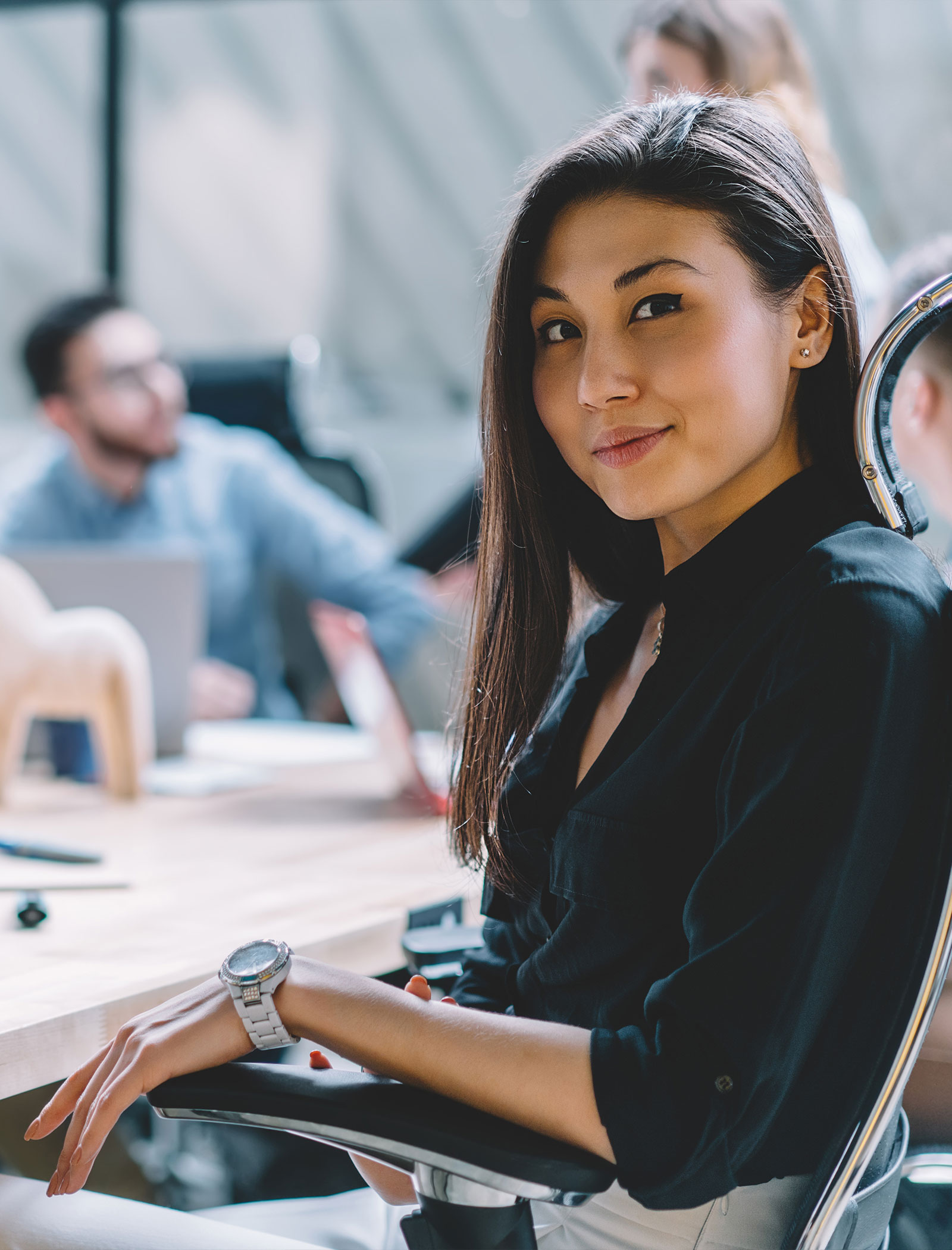 Young woman in a black shirt sitting on an office chair and looking confidently at the camera in a modern office.