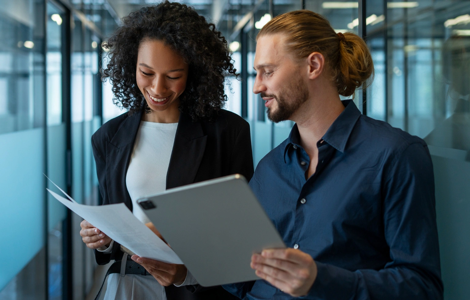Two coworkers review documents and a tablet together in a modern office corridor.