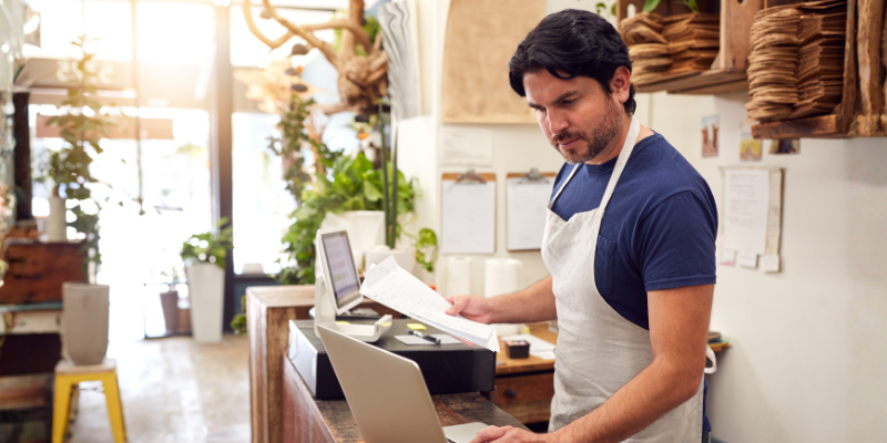 Male florist shop owner holding documents and looking at laptop