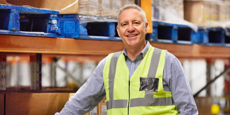 Import/export worker smiling, leaning on inventory and wearing bright yellow vest