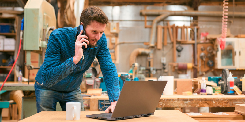 New Zealand carpenter working in woodwork workshop talking on mobile phone and using laptop