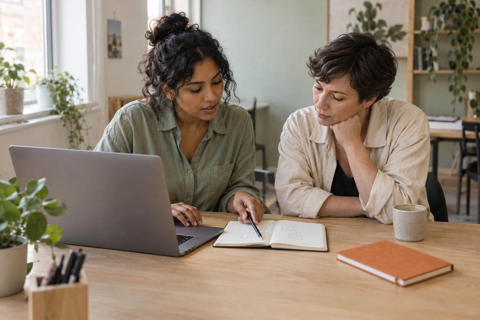 Two women reviewing a project diagram in a notebook beside a laptop at a shared workspace.
